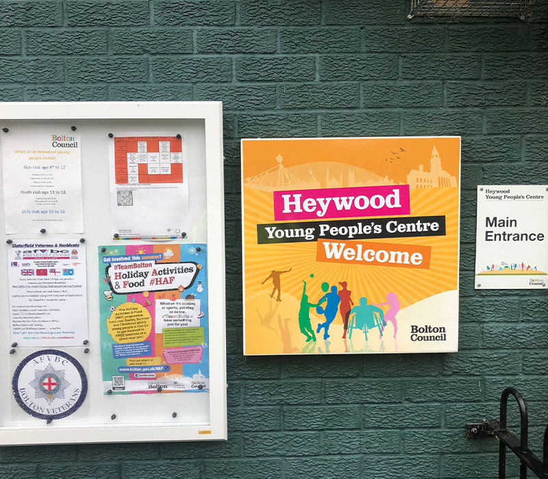 A Heywood Young Peoples Centre welcome sign with their yellow and orange branding, next to a notice board and main entrance sign on a green brick wall at the entrance to the Centre