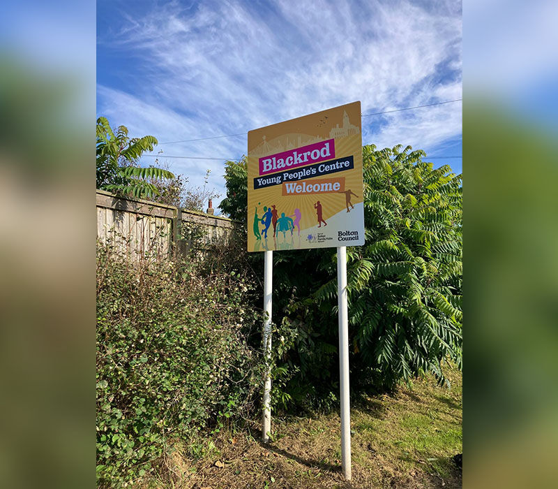 Wayfinding signage on the side of a road for Blackrod Young Peoples Centre, with their yellow branding