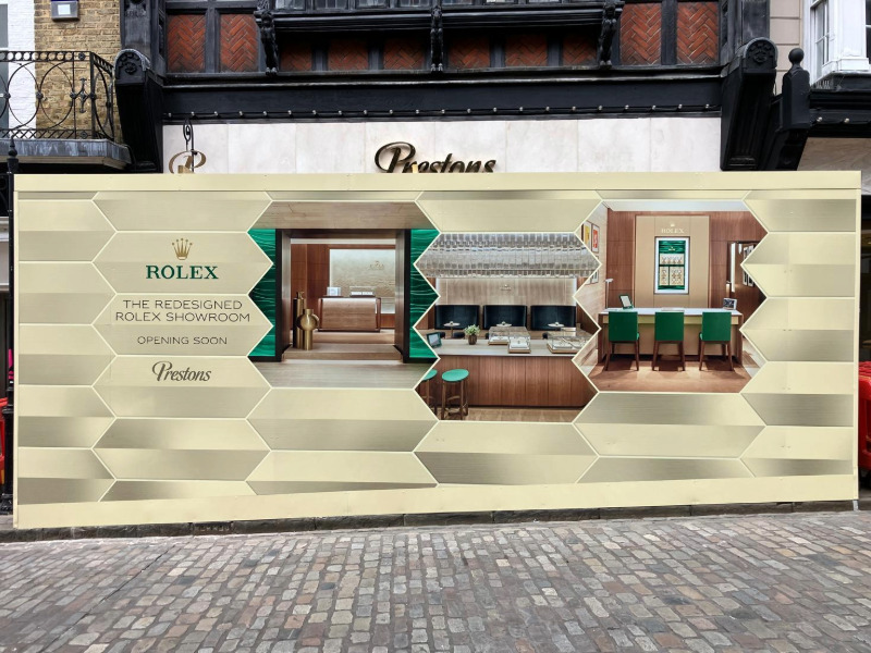 Construction hoarding covering Prestons Jewellers’ luxury showroom on a high street in Guildford. Hoarding says “The redesigned Rolex showroom. Opening soon.“
