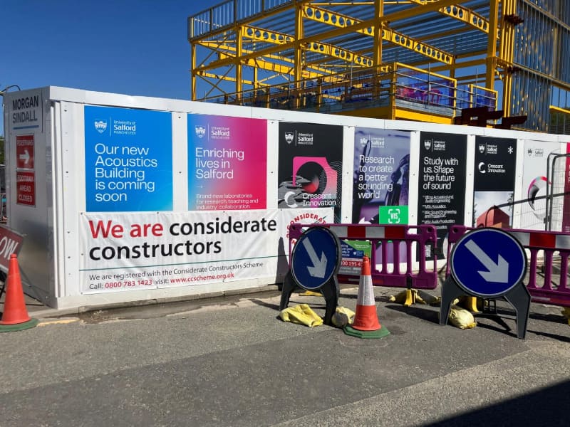 Construction hoarding for a new Acoustics Building at the University of Manchester. A yellow steel structure in the background under construction.